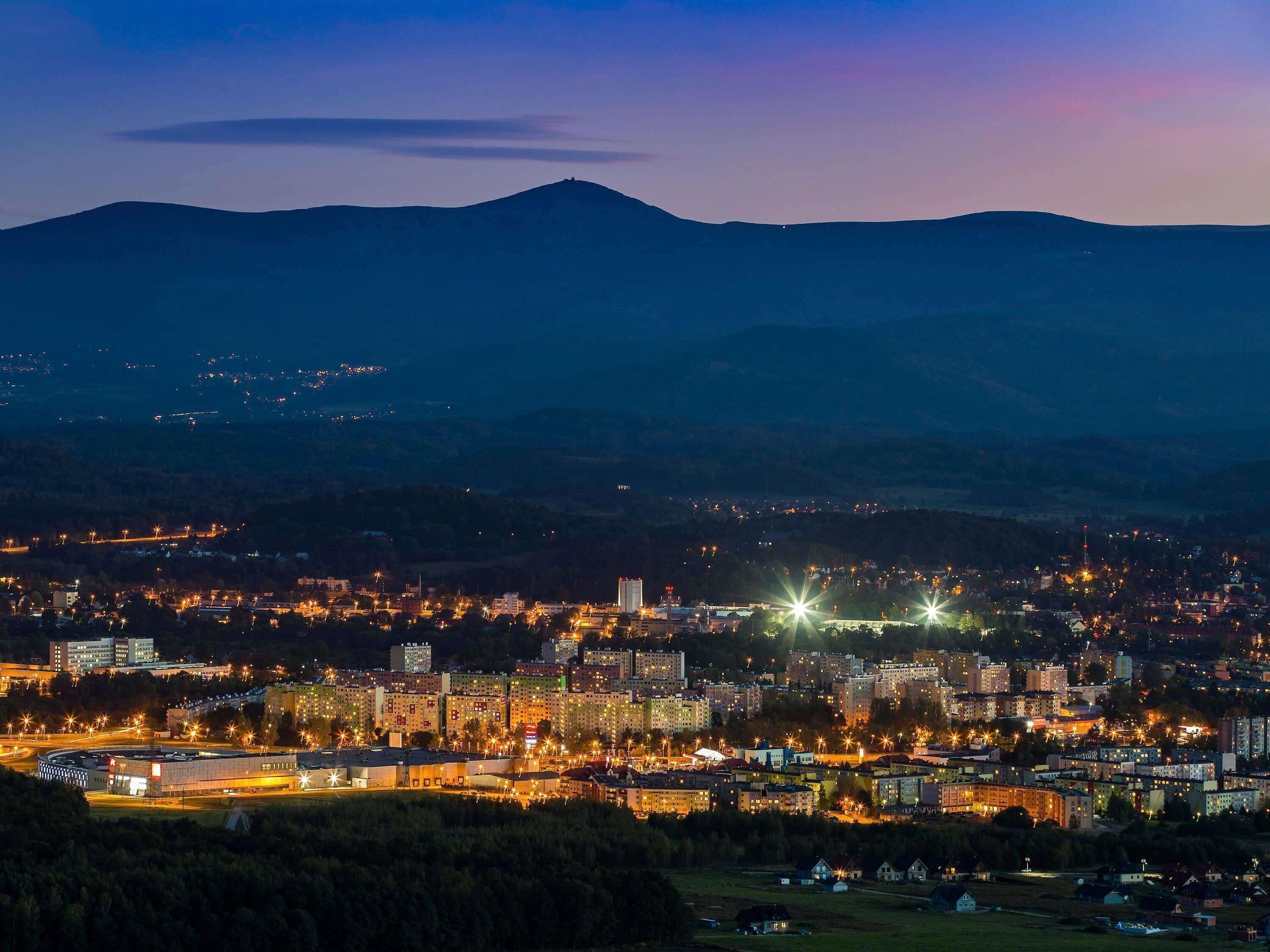 A cityscape at dusk with illuminated buildings and a mountain in the background.