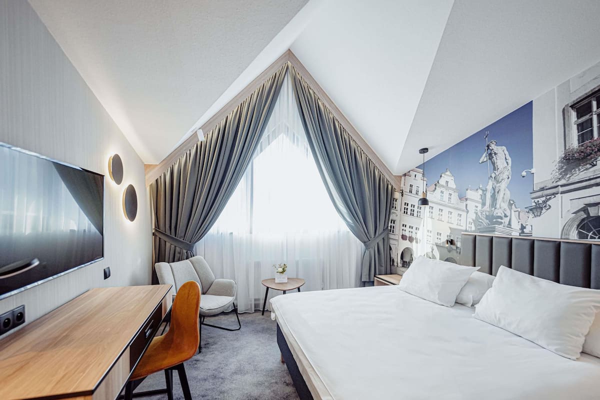 Ein modernes Hotelzimmer mit einem großen Bett, einem Schreibtisch, einem Sessel, einem Fernseher und Blick auf eine Stadtansicht durch das Fenster.