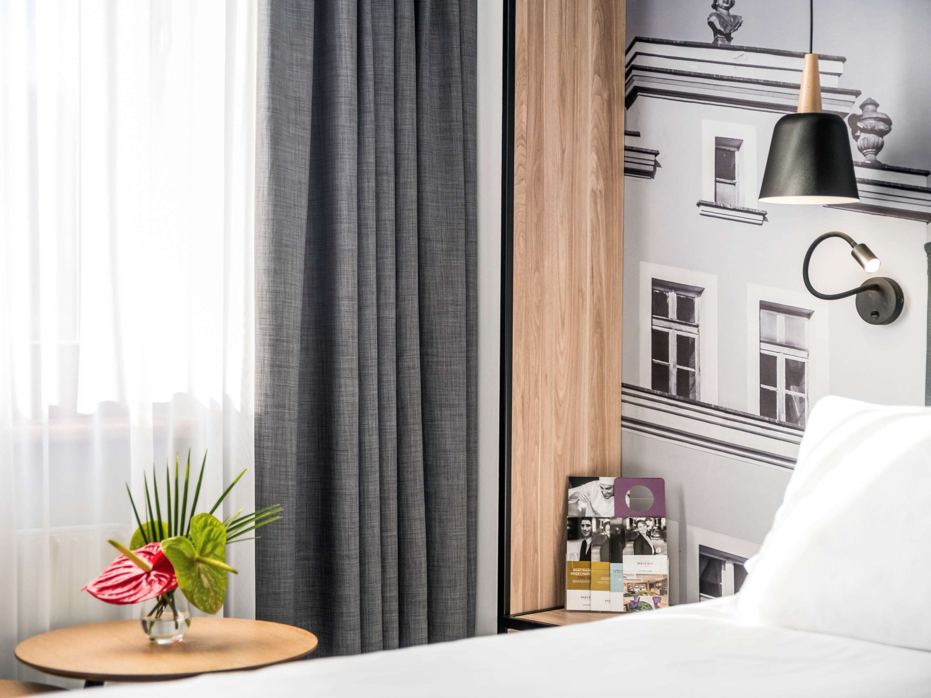Habitación de hotel con cama, una mesita redonda de madera con un jarrón de flores, una lámpara colgante negra y una pared con un collage de fotografías en blanco y negro.