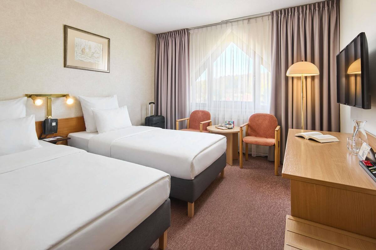 Ein Hotelzimmer mit zwei Betten, einem Schreibtisch, einem Stuhl, einem Fernseher, einer Lampe und einem fenster mit Vorhängen.