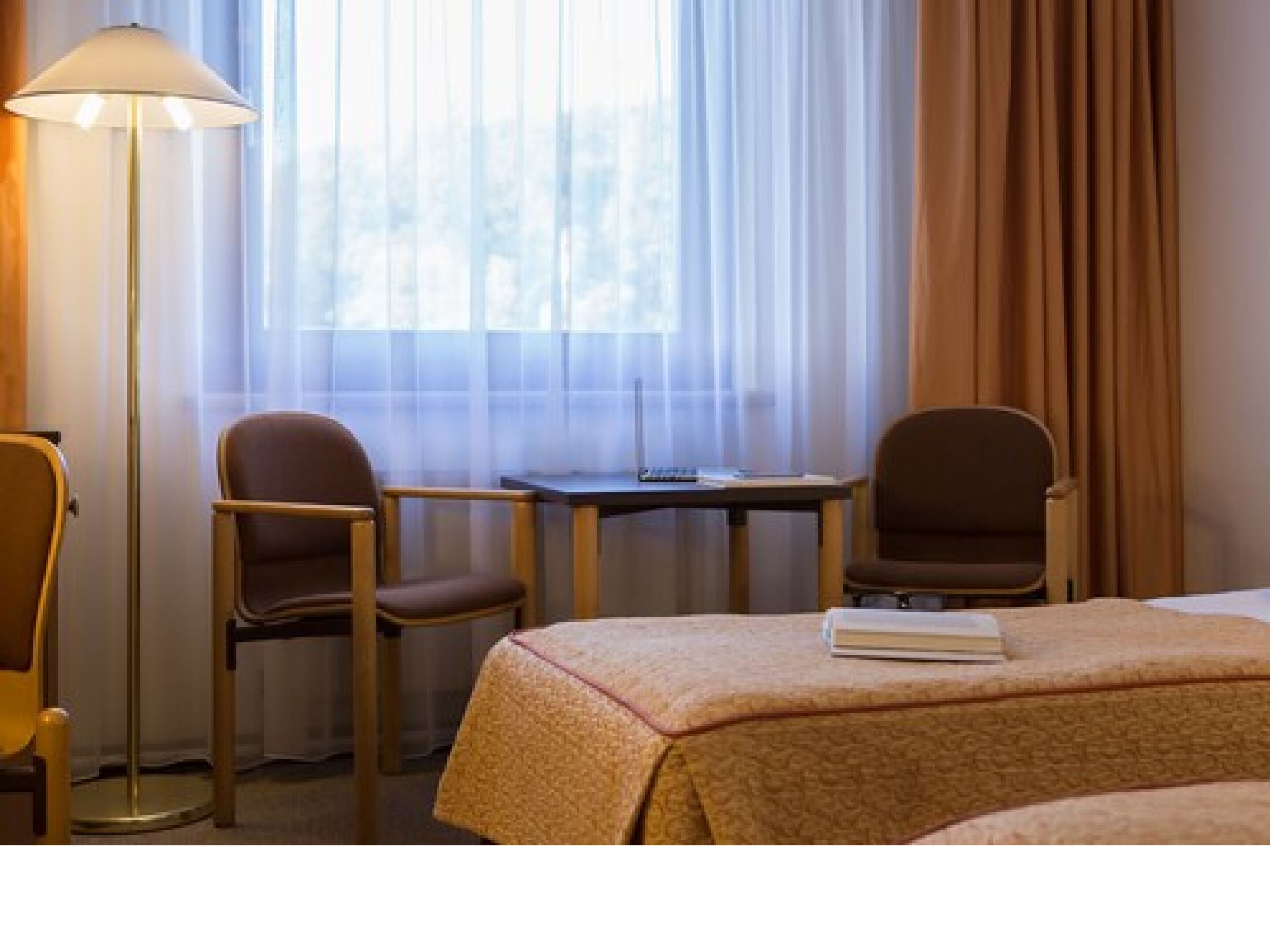 A hotel room with two chairs, a small table, a lamp, and a bed with a book on it.