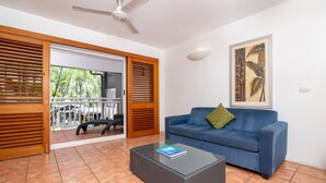 A living room with a blue sofa, a glass coffee table, and a ceiling fan. - Paradise on the Beach Resort - Palm Cove (Palm Cove)