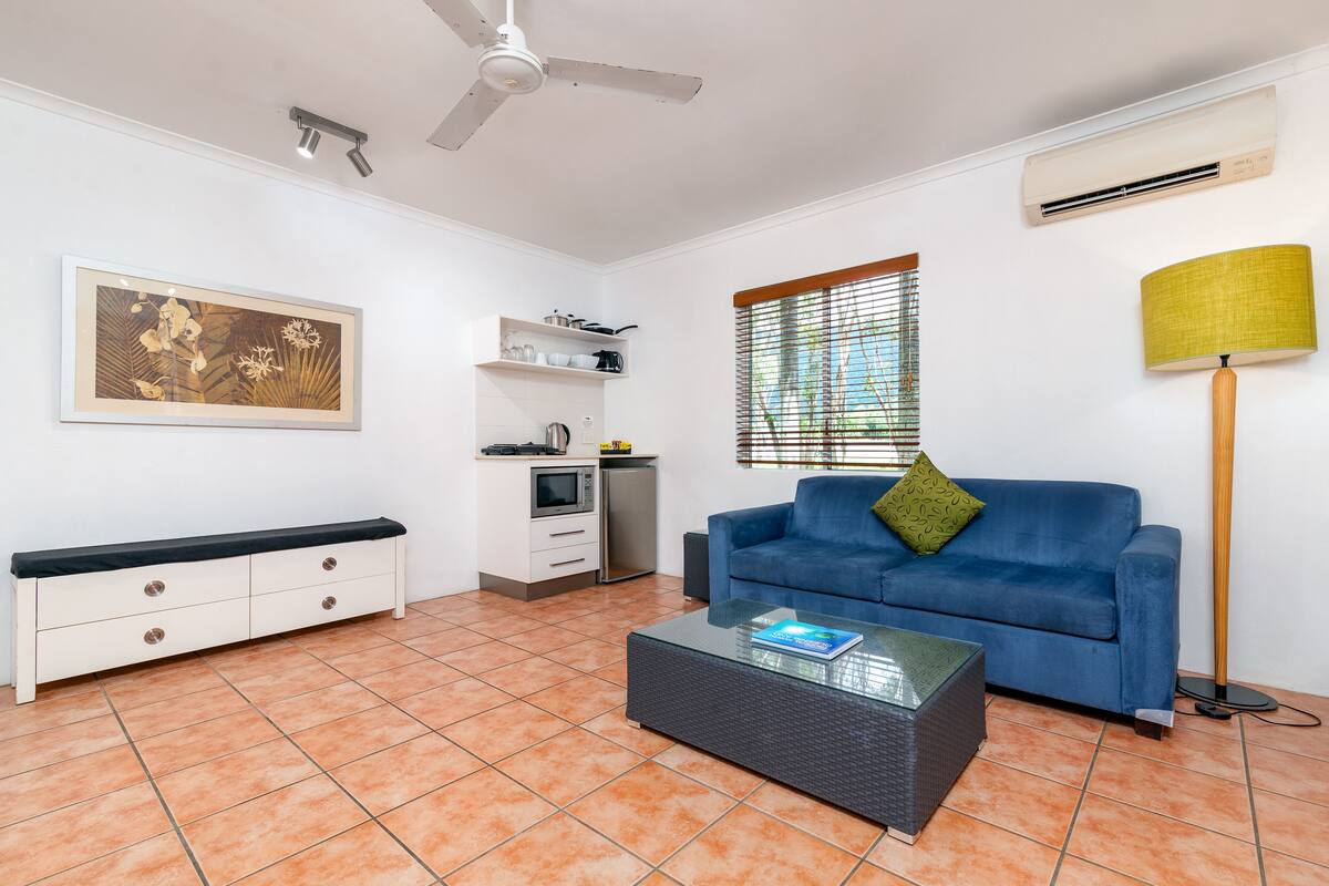 Ein Wohnzimmer mit blauem Sofa, Couchtisch, weißem Schrank, Deckenventilator und einem Fenster mit Jalousien.