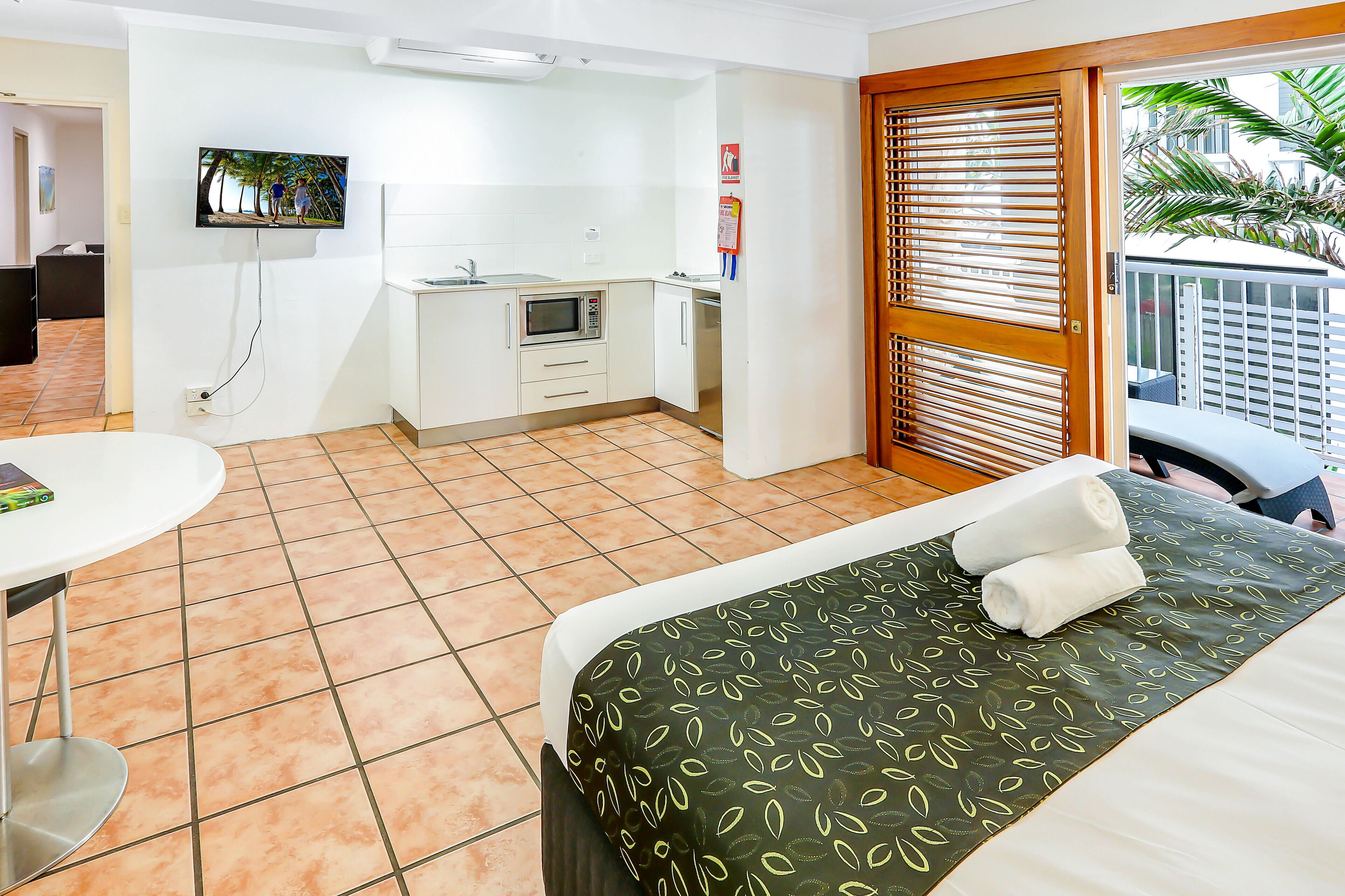 A hotel room with a bed, a small kitchenette, a TV mounted on the wall, and a balcony with a view of palm trees.