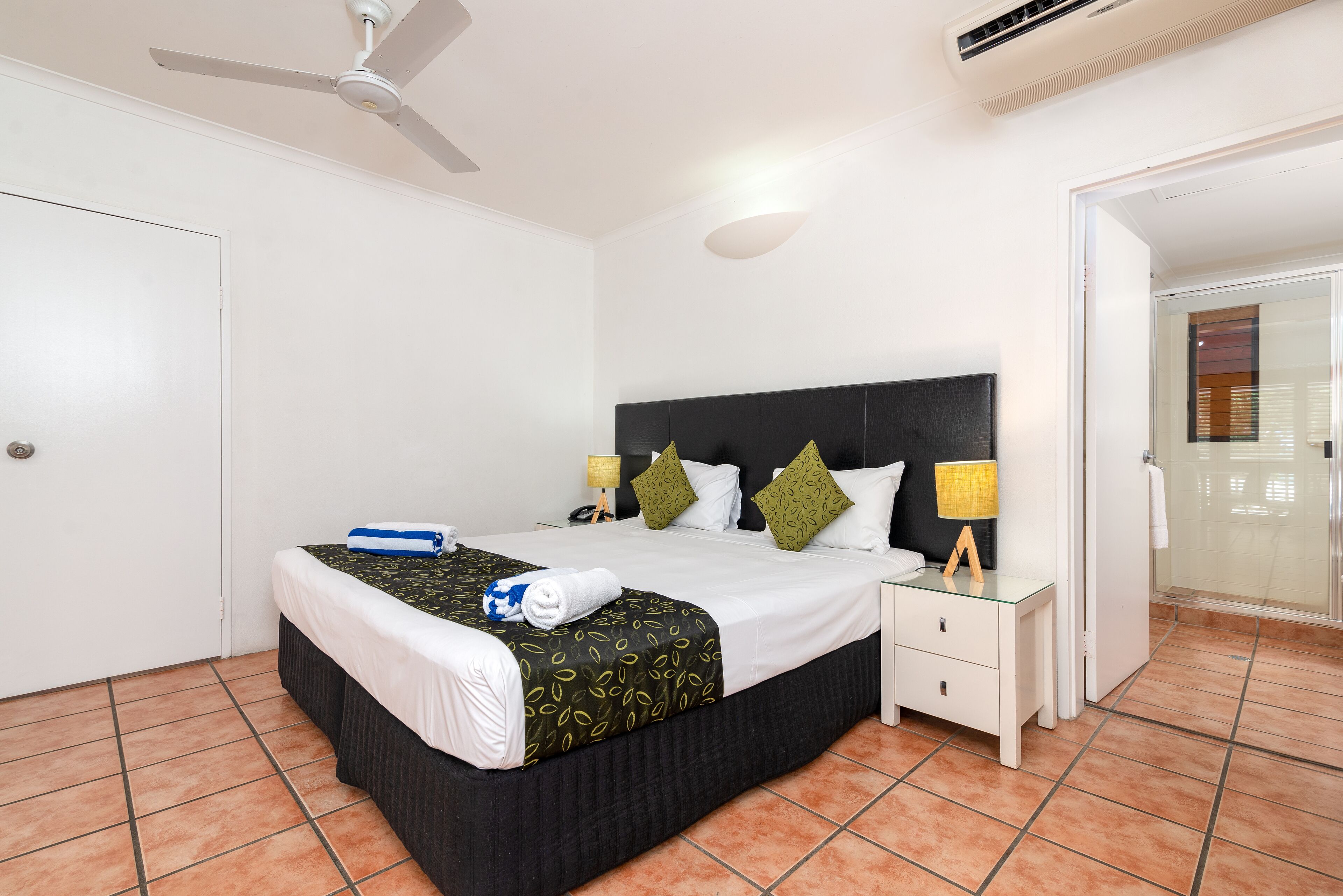 A neatly made bed with a black headboard, a bedside table with a lamp, and a bathroom visible through an open door.