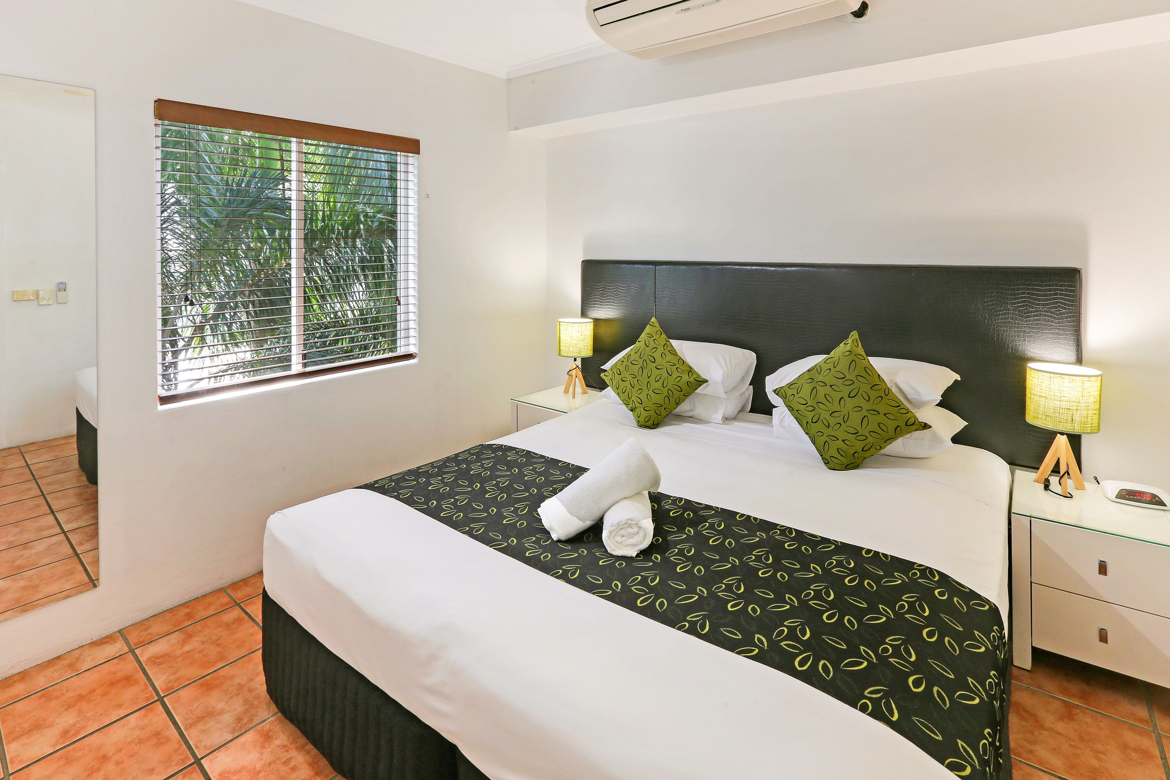 Una cama bien hecha con una colcha de diseño blanco y negro, dos almohadas verdes y una toalla blanca enrollada. Hay dos lámparas de noche con bases de madera, una mesita de noche pequeña y una ventana con persianas.
