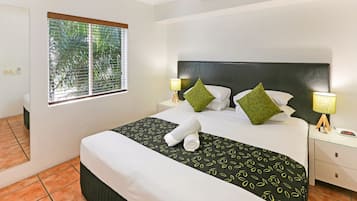 A neatly made bed with a black and white patterned bedspread, two green pillows, and a rolled white towel. There are two bedside lamps with wooden bases, a small bedside table, and a window with blinds.