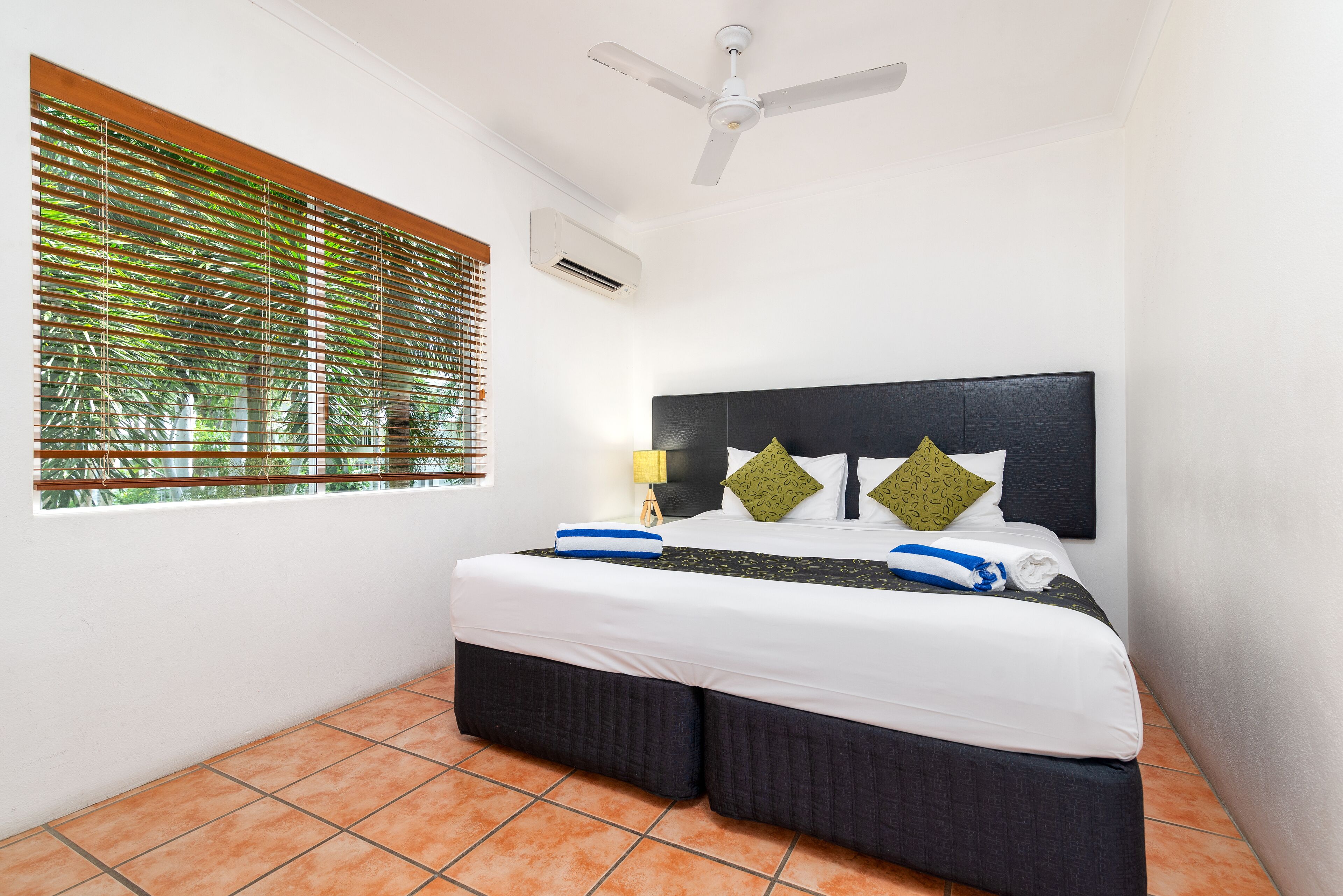 A hotel room with a bed, a ceiling fan, an air conditioning unit, and a window with blinds.