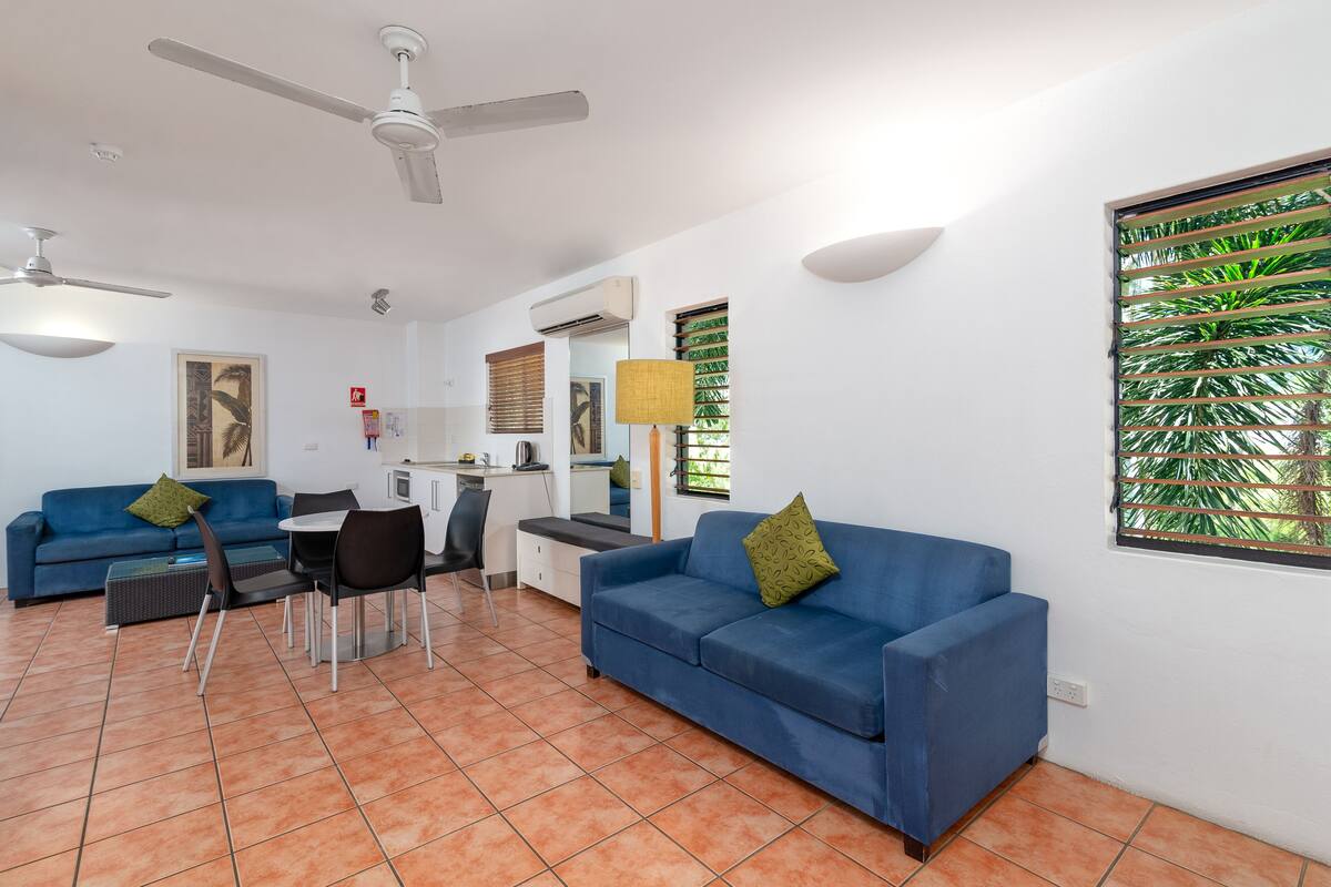 Ein modernes Wohnzimmer mit einem blauen Sofa, einem Esstisch, Stühlen, Deckenventilatoren und einem Fenster mit Jalousien.
