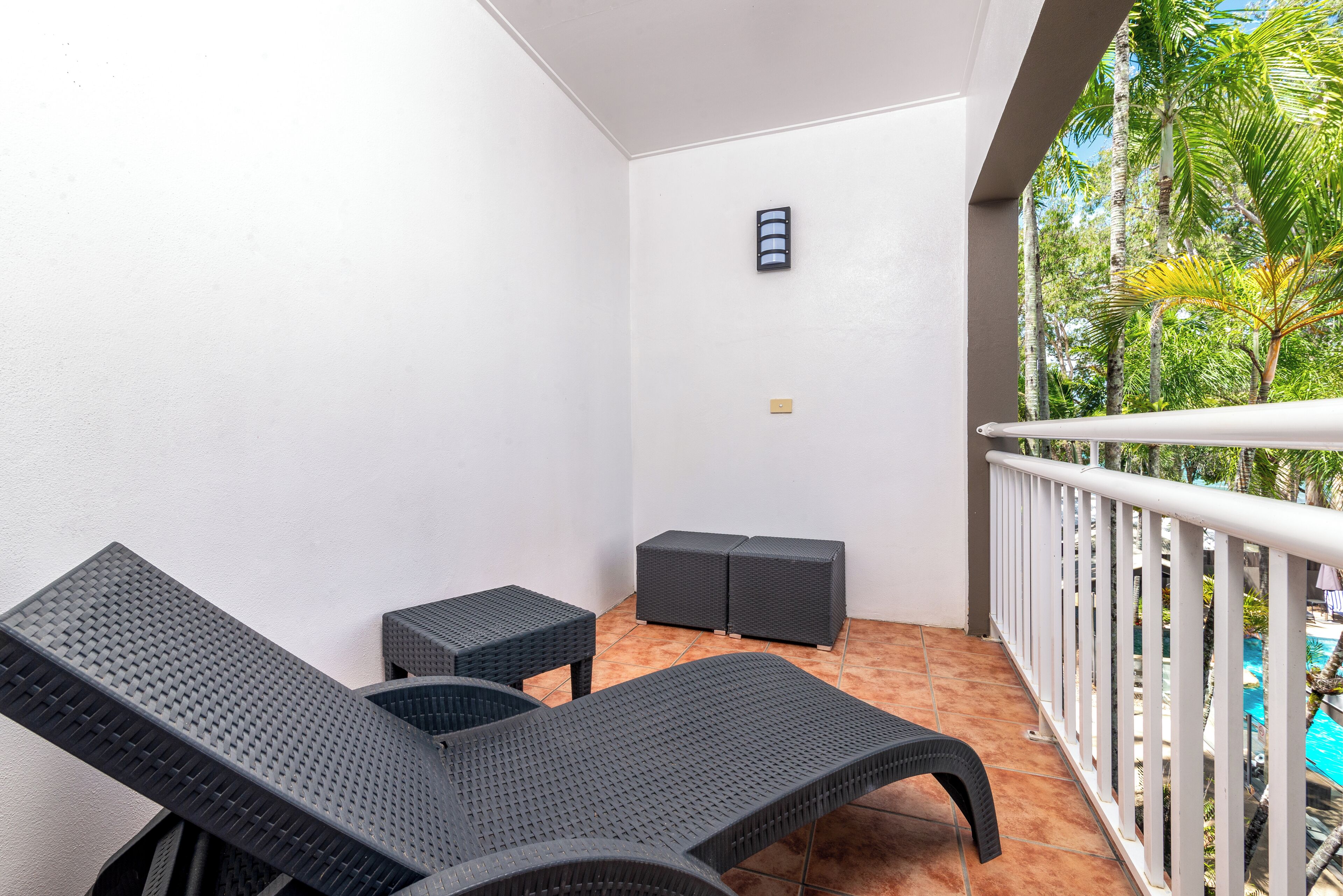 A balcony with two lounge chairs and a small table, a white railing, and a view of a swimming pool.