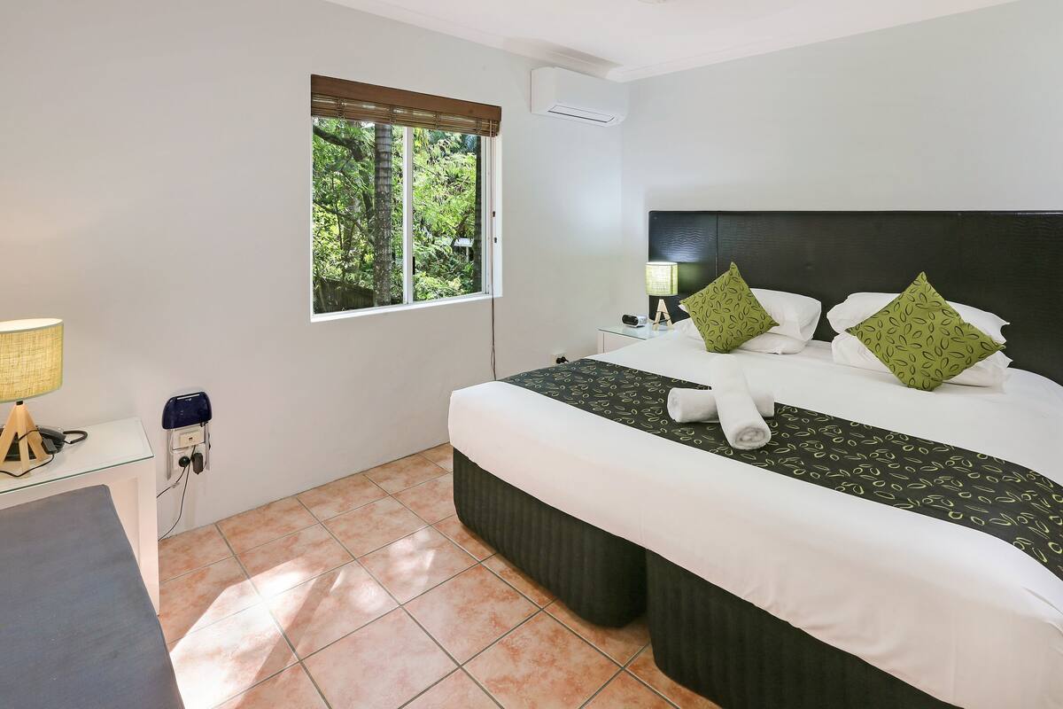 Ein Hotelzimmer mit Bett, Schreibtisch, Lampe, einem Fenster mit Jalousien und einer Klimaanlage.
