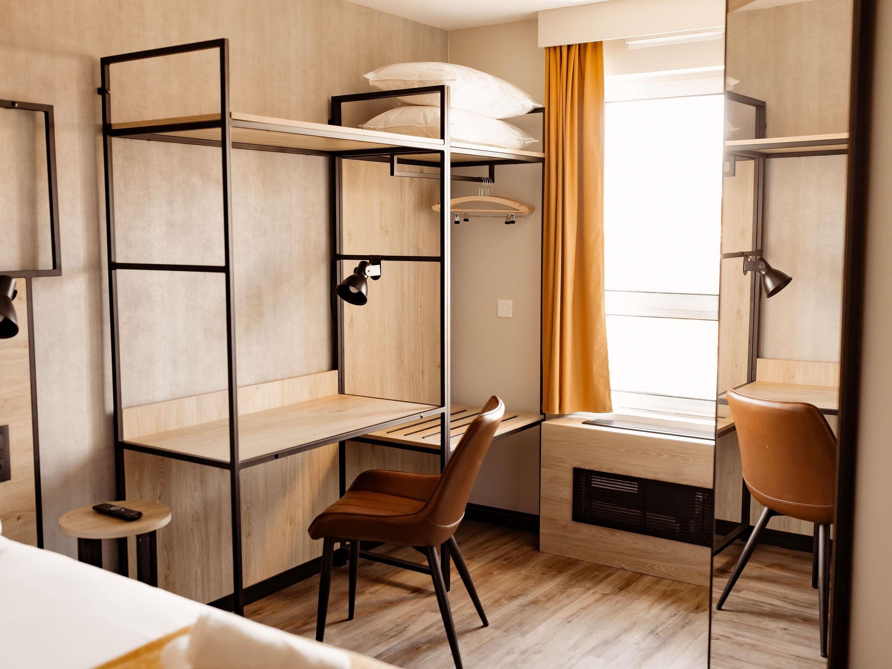 A compact hotel room with a bunk bed, a desk, a chair, a small table, and a window with curtains.