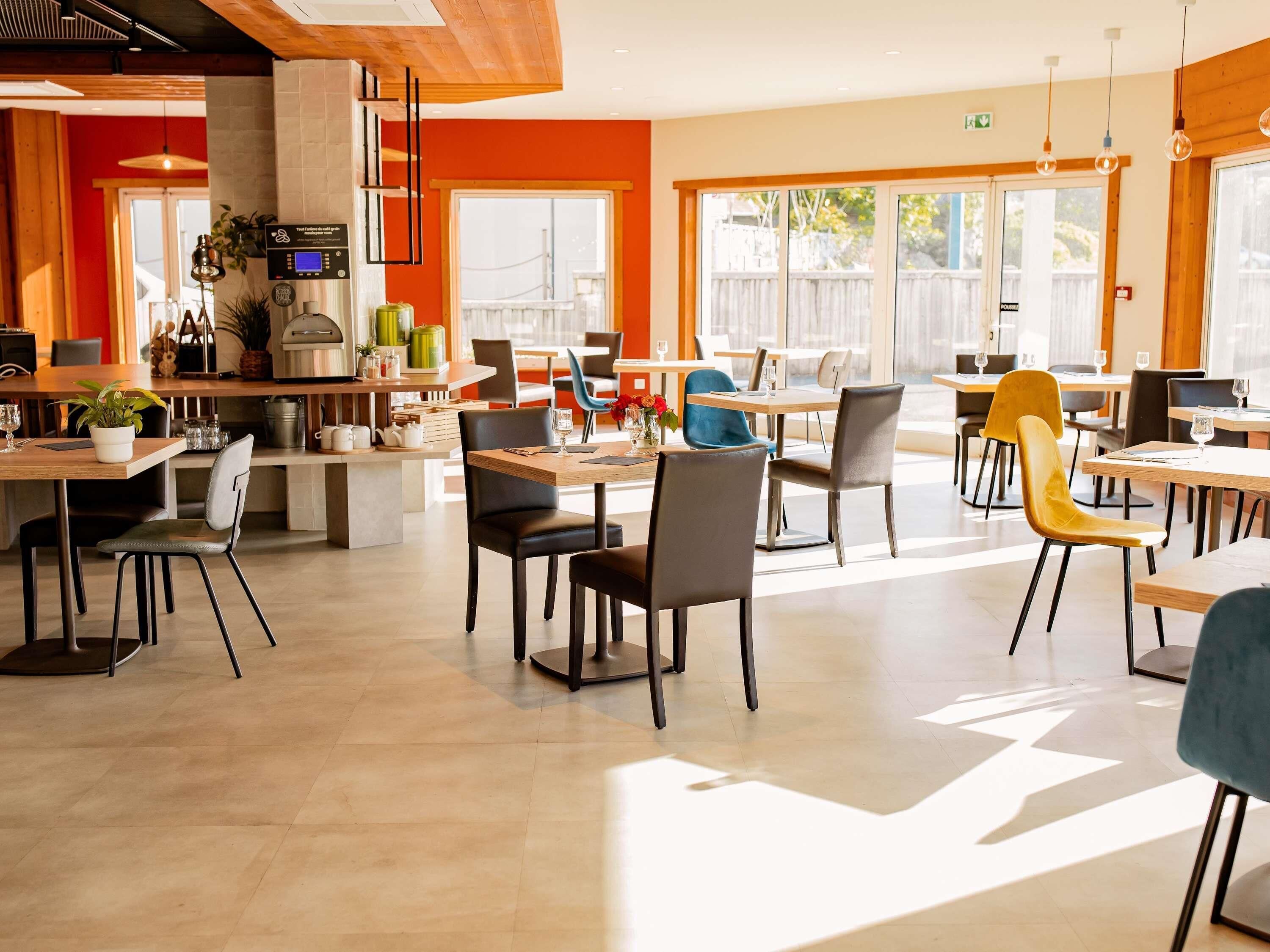 Un moderno comedor con varias mesas y sillas, una zona de cocina con electrodomésticos, y grandes ventanales que dejan entrar luz natural.
