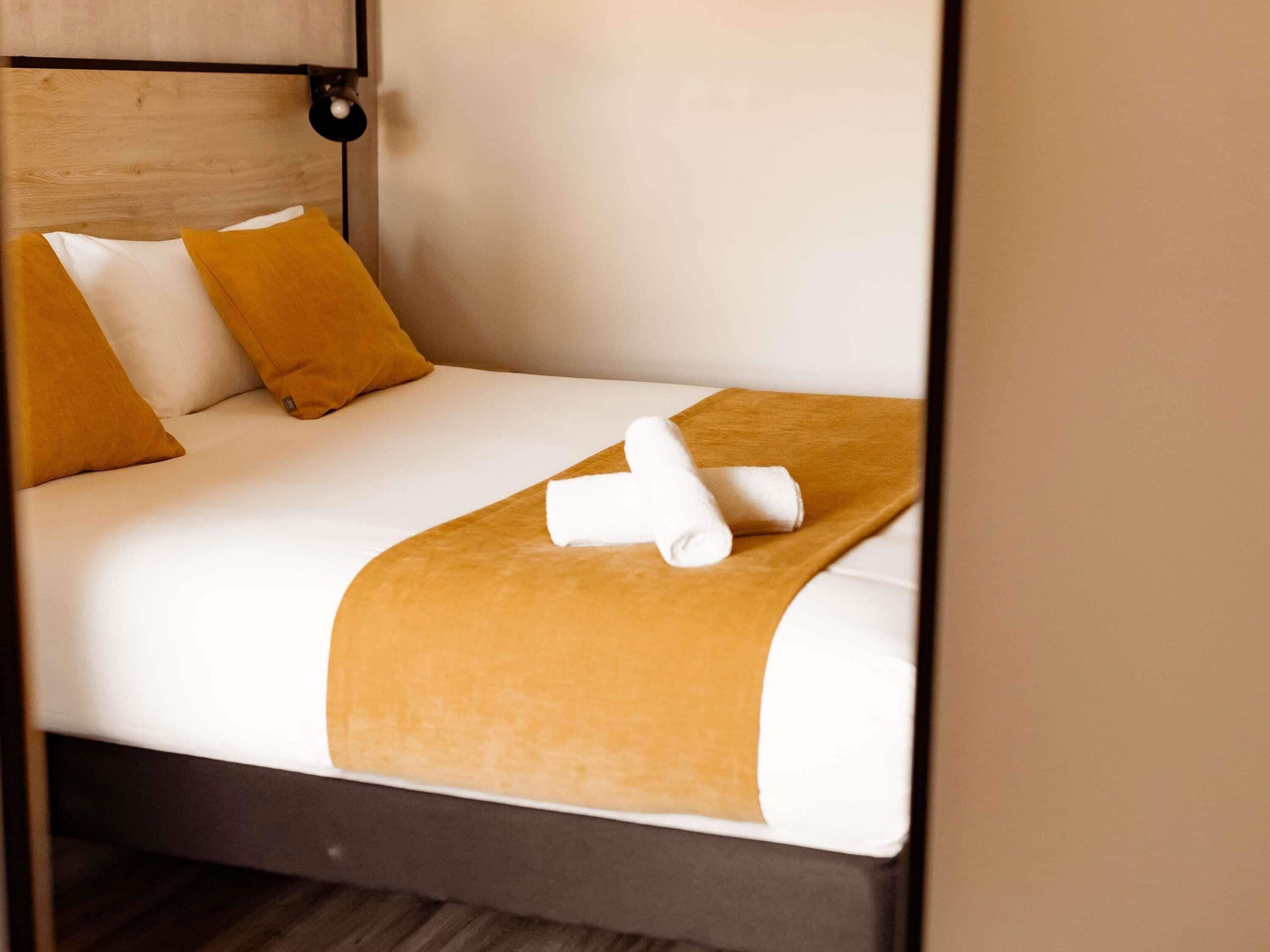 Una cama bien hecha con ropa blanca, dos almohadas color mostaza y una toalla blanca enrollada sobre una manta marrón.