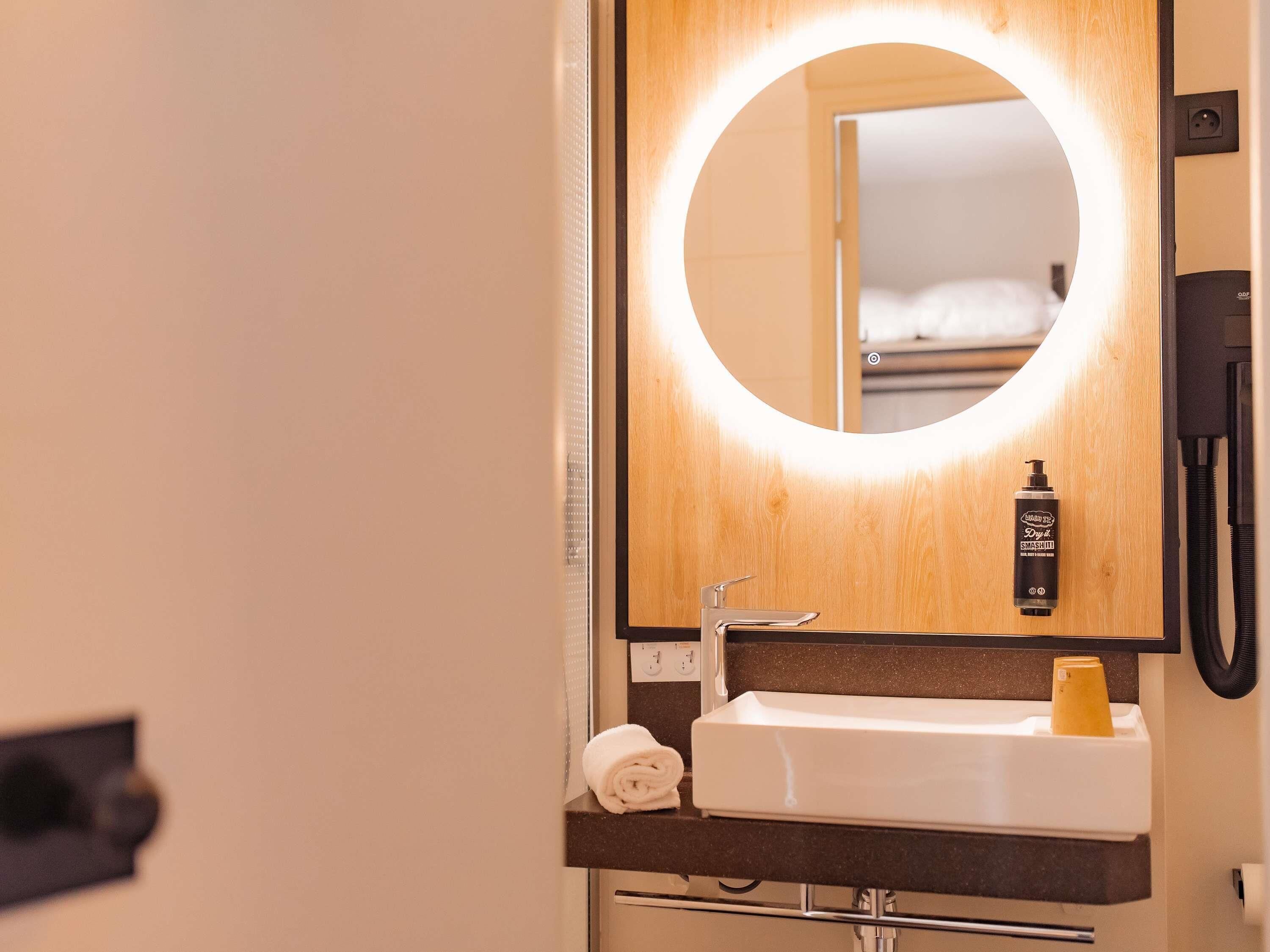 Un baño de hotel con un espejo redondo, un lavabo, una toalla y una botella de champú.