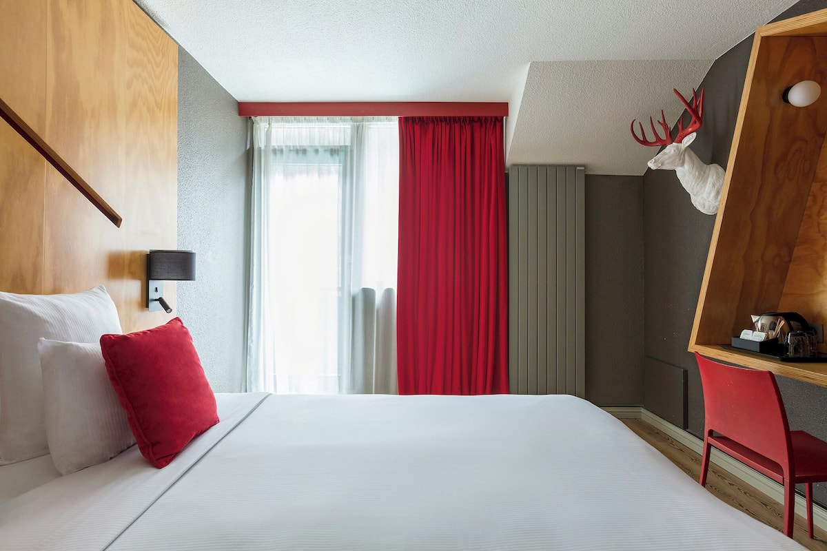 Een hotelkamer met een bed, een rode stoel, een houten bureau, een rood gordijn en een op de muur bevestigd hertenhoofd.
