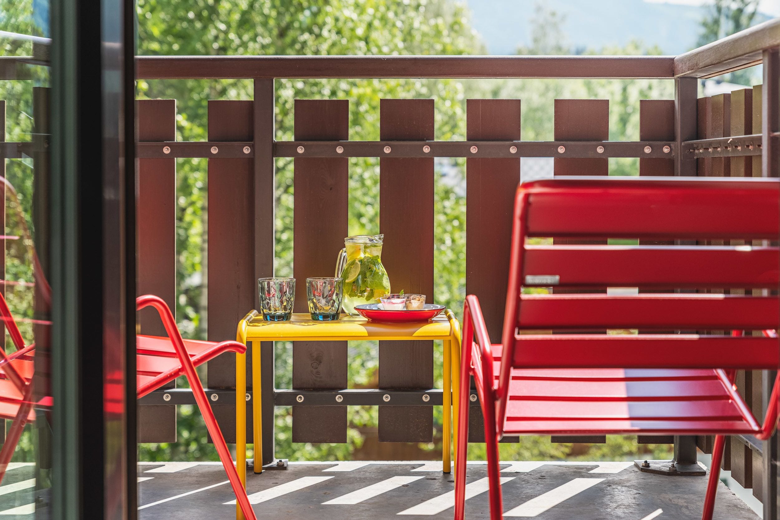 A balcony with a yellow table, red chairs, and a glass of iced tea.