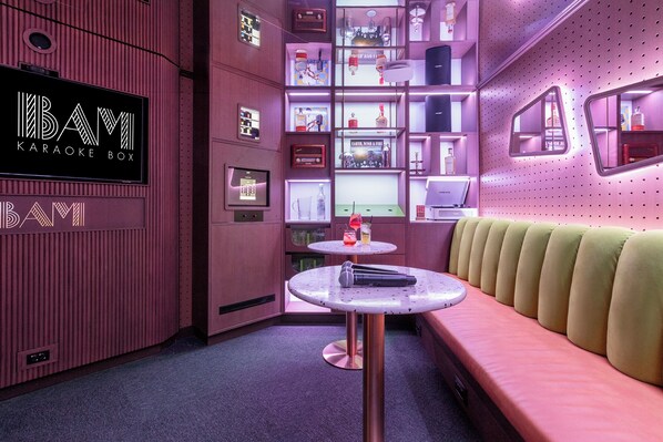 A karaoke room with a pink and purple color scheme, a karaoke box sign, shelves with various items, and a seating area with a table and microphone. - Hôtel Les Aiglons Chamonix  (Chamonix-Mont-Blanc)