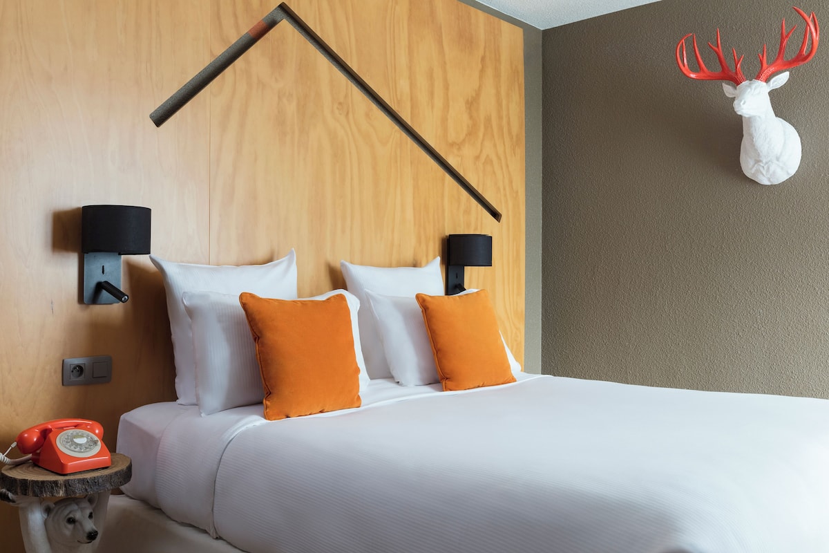 Hotelkamer met een houten hoofdeinde, witte beddengoed, twee oranje kussens, een opgehangen hertenkop en een klassieke rode draaischijf telefoon op een klein tafeltje.