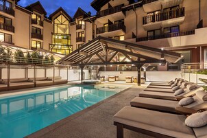 Uma área junto à piscina com espreguiçadeiras e um espaço de estar coberto, cercada por um edifício com varandas e grandes janelas.