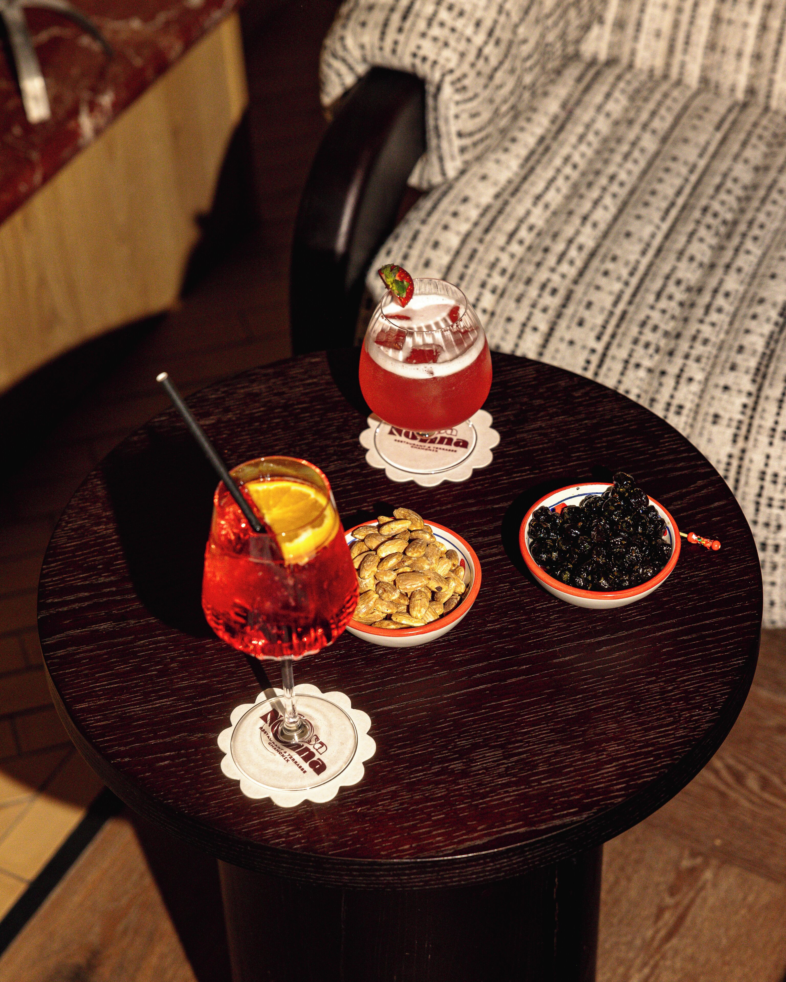 Una mesa redonda con superficie de madera oscura, un vaso de bebida roja con una rodaja de limón y una pajita, un cuenco de almendras y un cuenco de aceitunas negras.