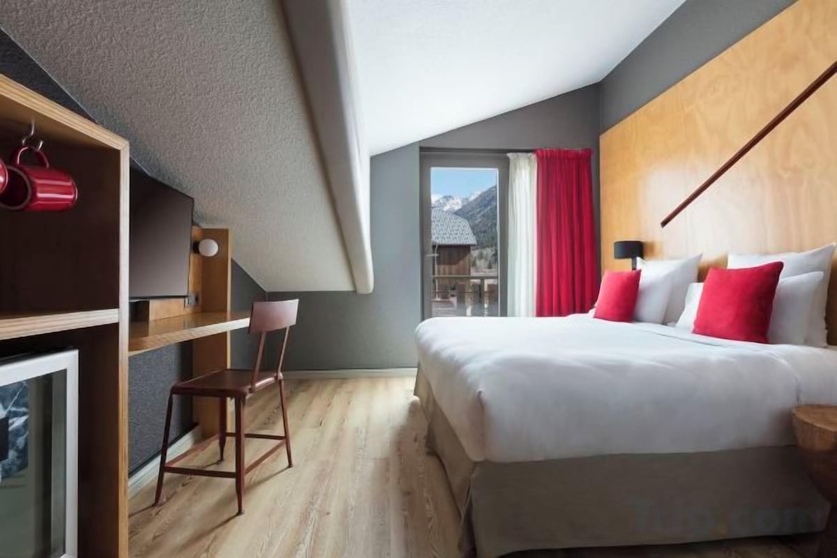 Une chambre d’hôtel moderne avec un grand lit, un bureau avec une chaise et une vue sur les montagnes par la fenêtre.