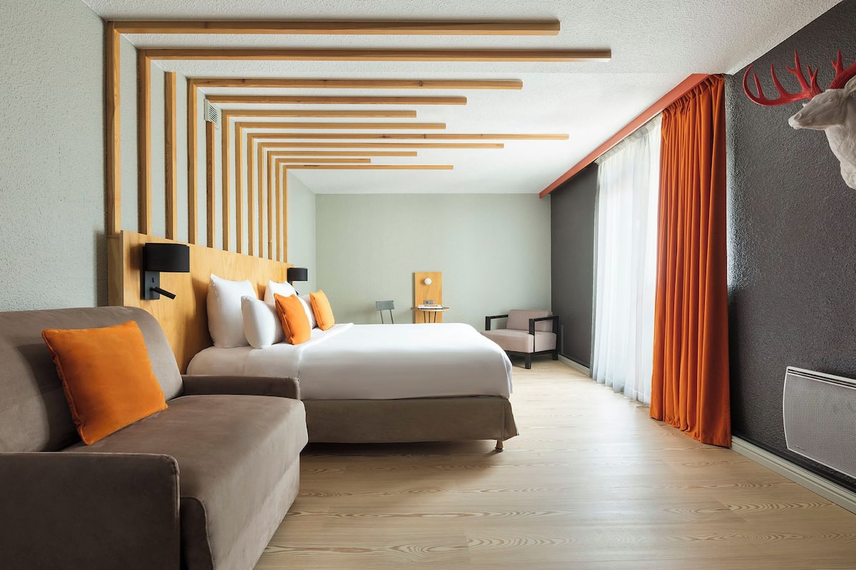 Een moderne hotelkamer met een bed, een bank, een bijzettafeltje en een groot raam met gordijnen.