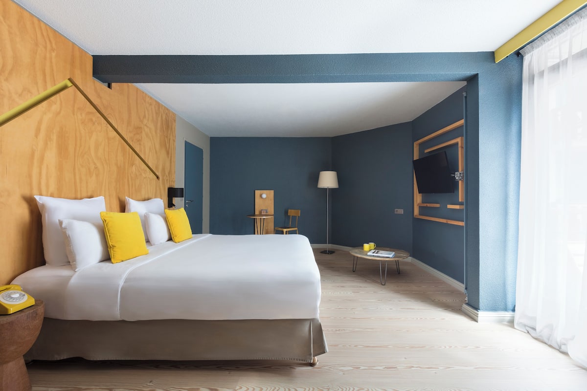 Een hotelkamer met een groot bed, een houten hoofdbord, een flatscreen televisie aan de muur, een tafeltje met een lamp en een raam met gordijnen.
