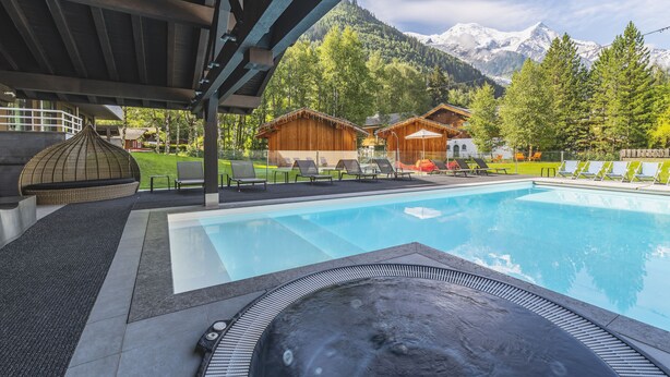 A swimming pool with a hot tub, lounge chairs, and wooden cabins with a mountain backdrop.