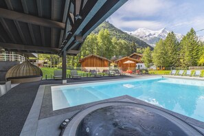 A swimming pool with a hot tub, lounge chairs, and wooden cabins with a mountain backdrop. - Hôtel Les Aiglons Chamonix  (Chamonix-Mont-Blanc)