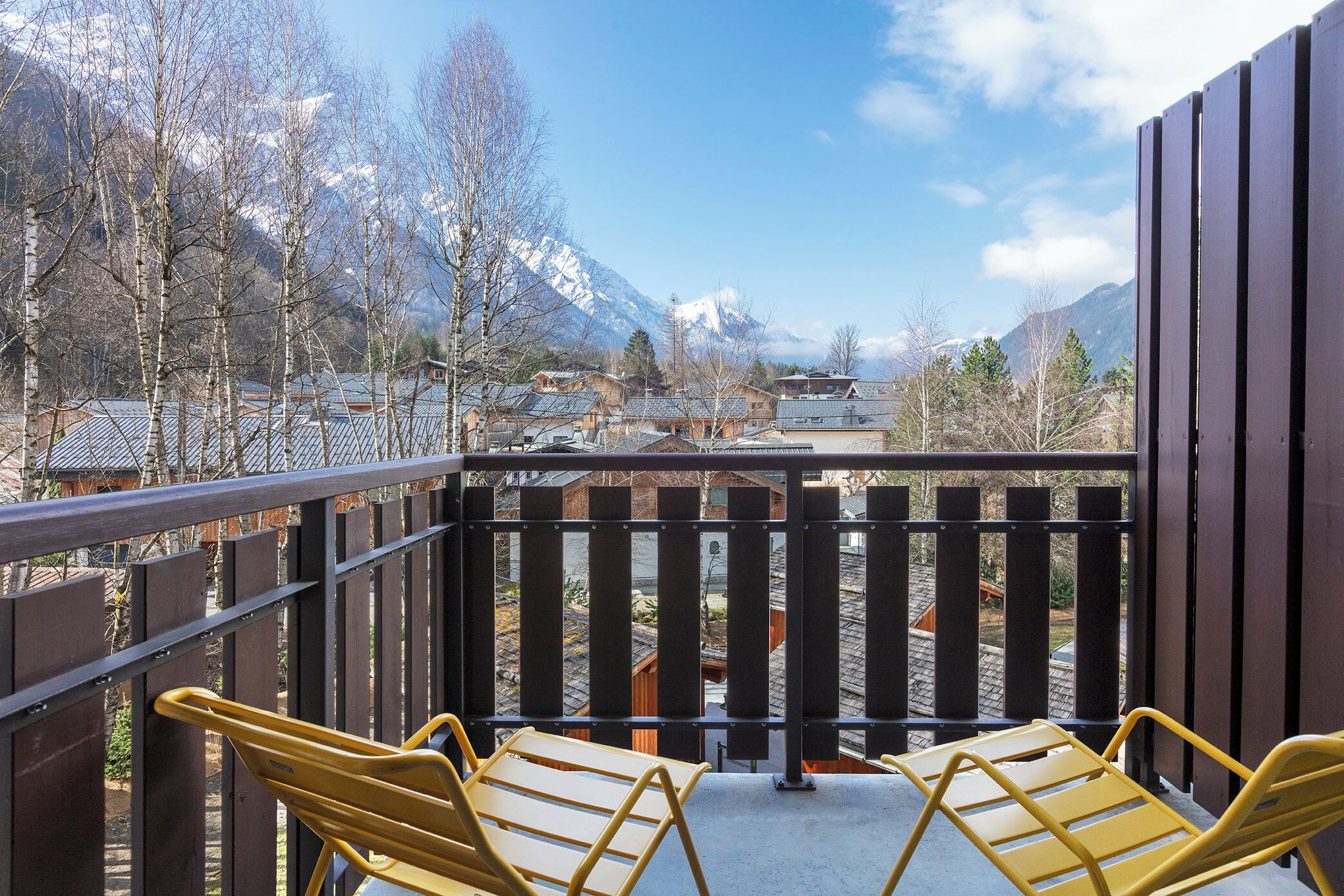 Een balkon met twee gele stoelen en uitzicht op besneeuwde bergen, met een houten hek.