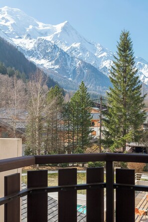 Une vue sur une montagne avec des sommets enneigés, une balustrade en bois sur un balcon et un bâtiment avec un toit en pente au premier plan.