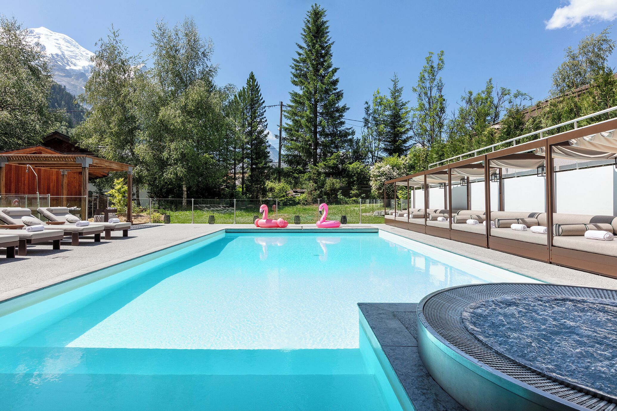 Una piscina con sillas de descanso y un jacuzzi, rodeada de árboles y una montaña al fondo.