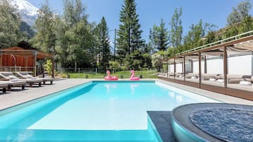 A swimming pool with lounge chairs and a hot tub, surrounded by trees and a mountain in the background.