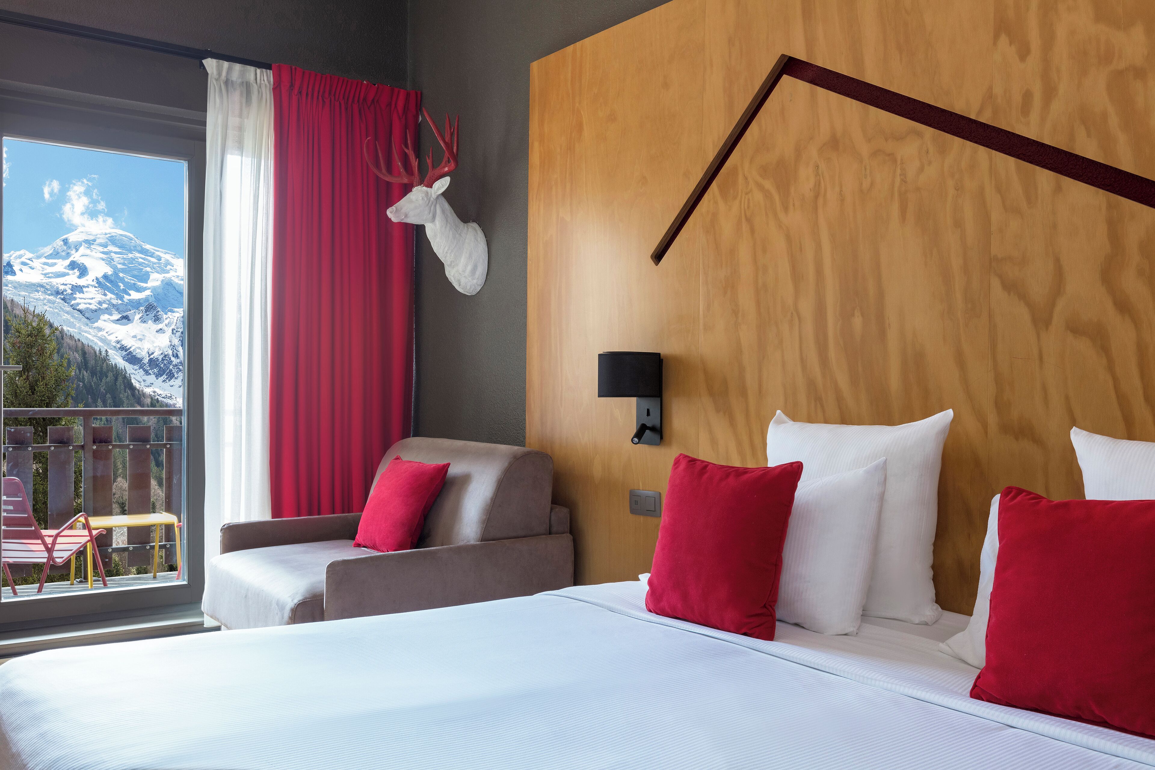 Ein Hotelzimmer mit einem großen Bett, zwei Sesseln, einem hölzernen Kopfteil, einem aufgehängten Hirschgeweih und Blick auf schneebedeckte Berge.