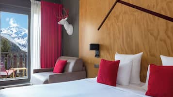 Ein Hotelzimmer mit einem großen Bett, zwei Sesseln, einem hölzernen Kopfteil, einem aufgehängten Hirschgeweih und Blick auf schneebedeckte Berge.