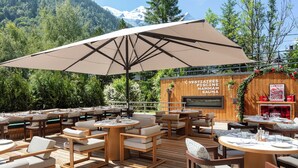 An outdoor seating area with wooden tables and chairs, a large umbrella, and a wooden wall with signage for a hammam sauna.