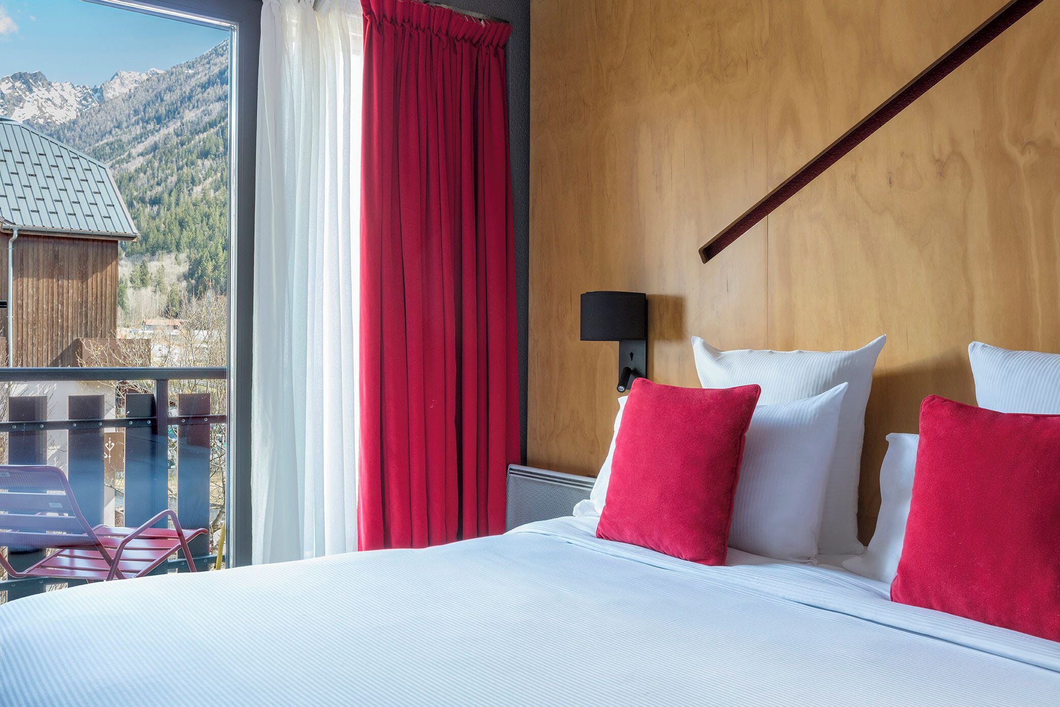 A hotel room with a large bed, red and white pillows, a wooden headboard, a nightstand with a lamp, and a view of mountains through a large window.