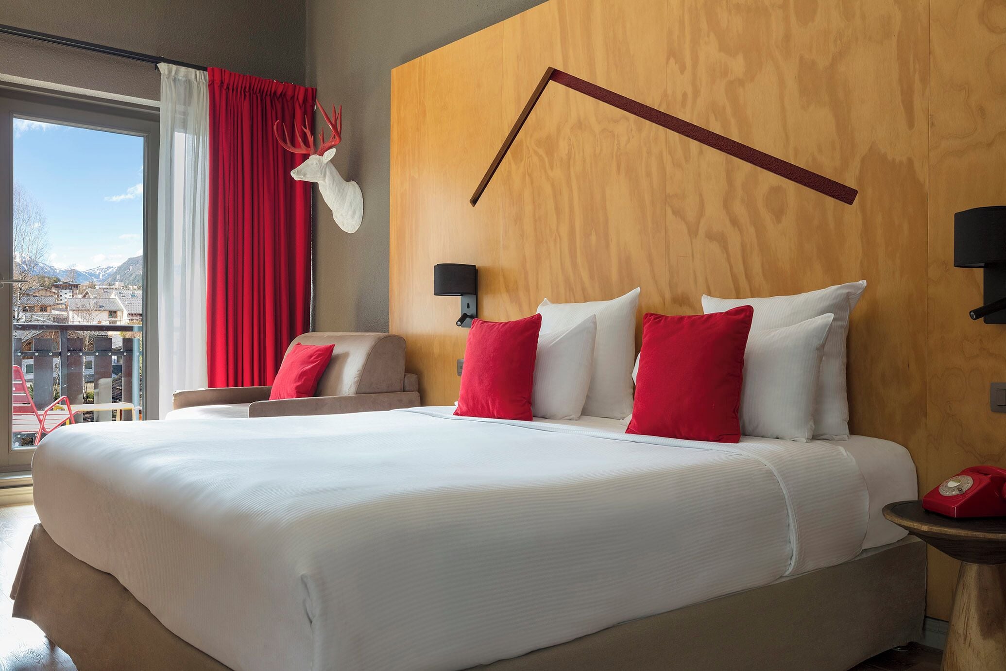 A hotel room with a large bed, red and white pillows, a wooden headboard, a nightstand with a red telephone, and a view of a cityscape through a large window.