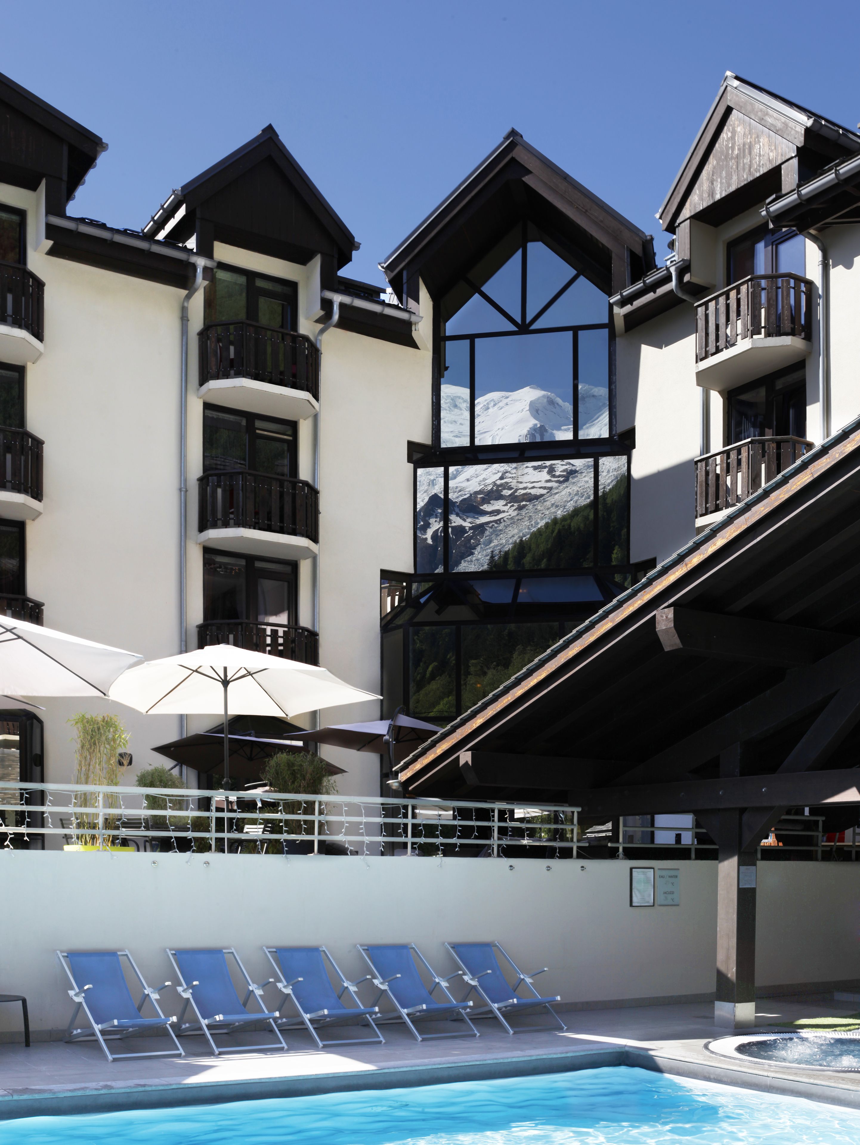 Un edificio moderno con balcones, una zona de piscina con tumbonas y vistas a una montaña.