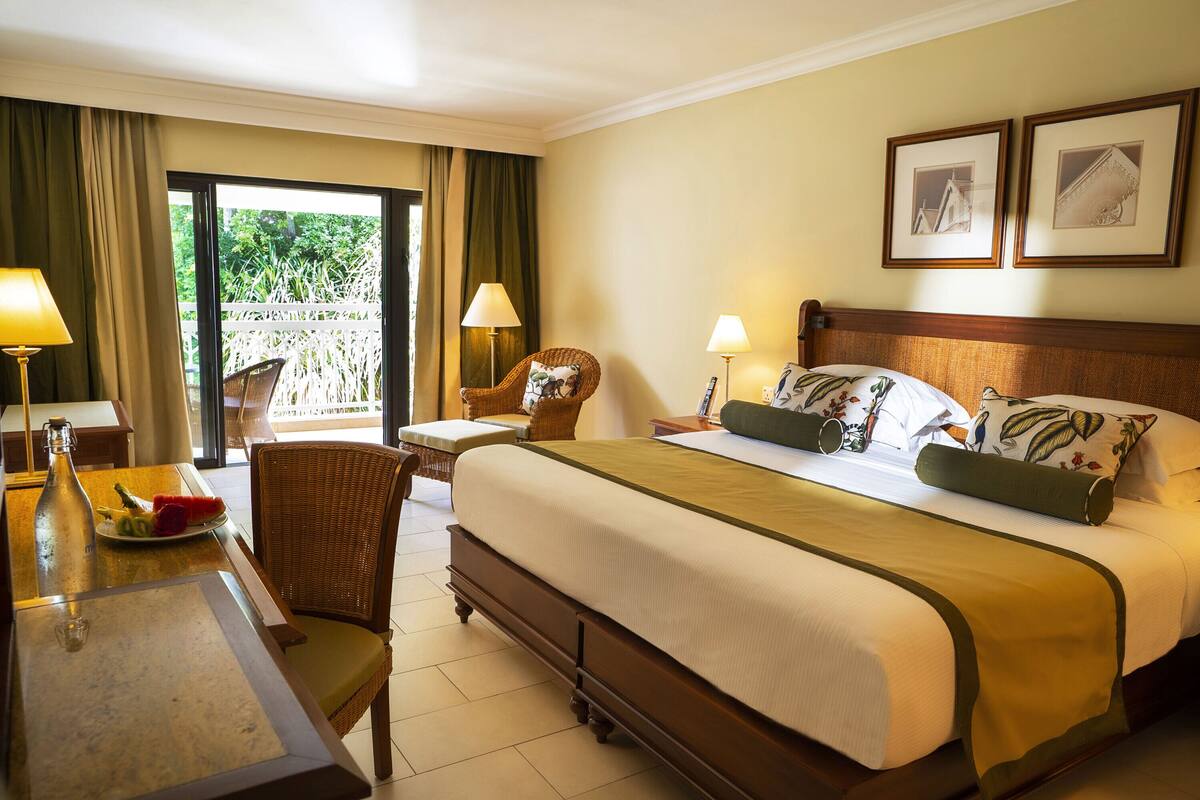 Ein Hotelzimmer mit einem großen Bett, zwei Nachttischlampen, einem Korbstuhl, einem Esstisch mit einer Flasche und Obst, sowie einem Blick auf Grün hinter dem Fenster.