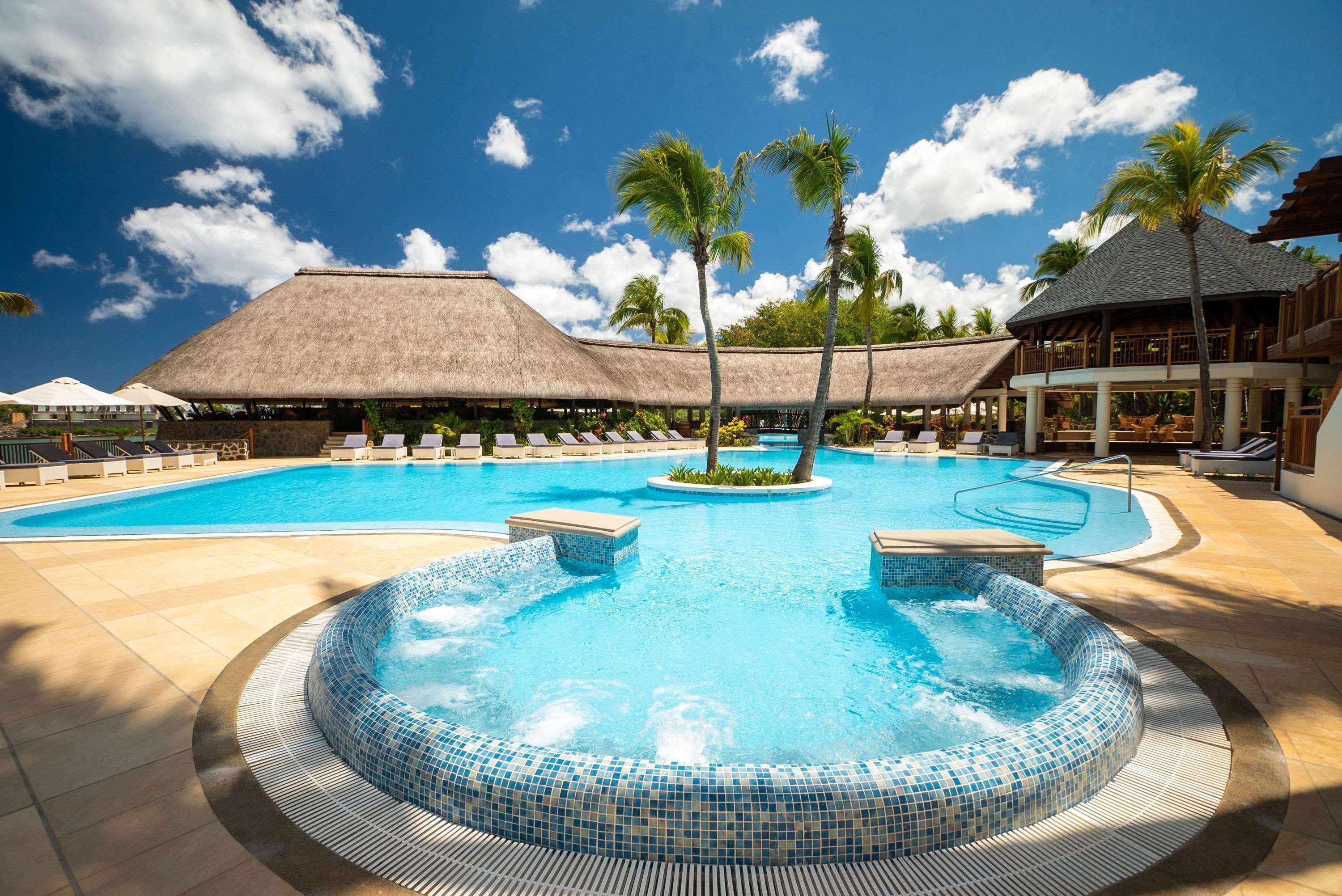 A resort pool with a hot tub, lounge chairs, and thatched-roof buildings.