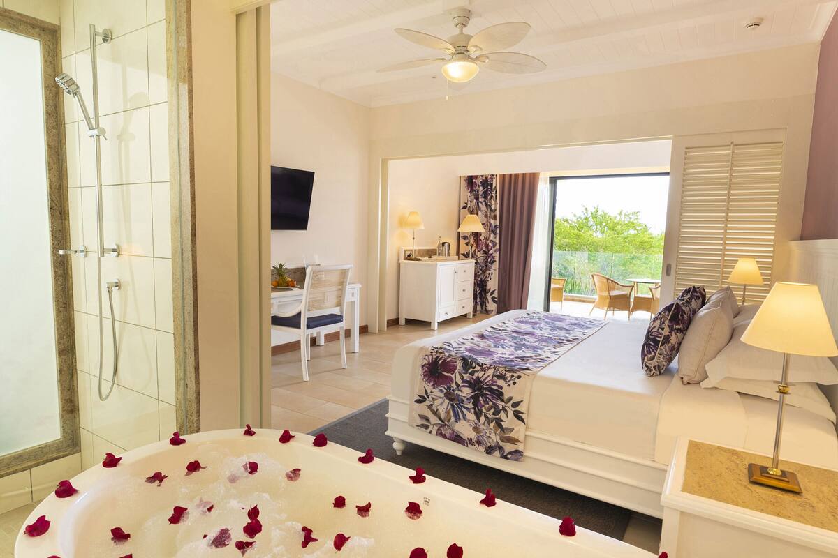 Ein Schlafzimmer mit einem großen Bett, einer Sitzecke mit Schreibtisch, einer Dusche und Blick auf Grünflächen.