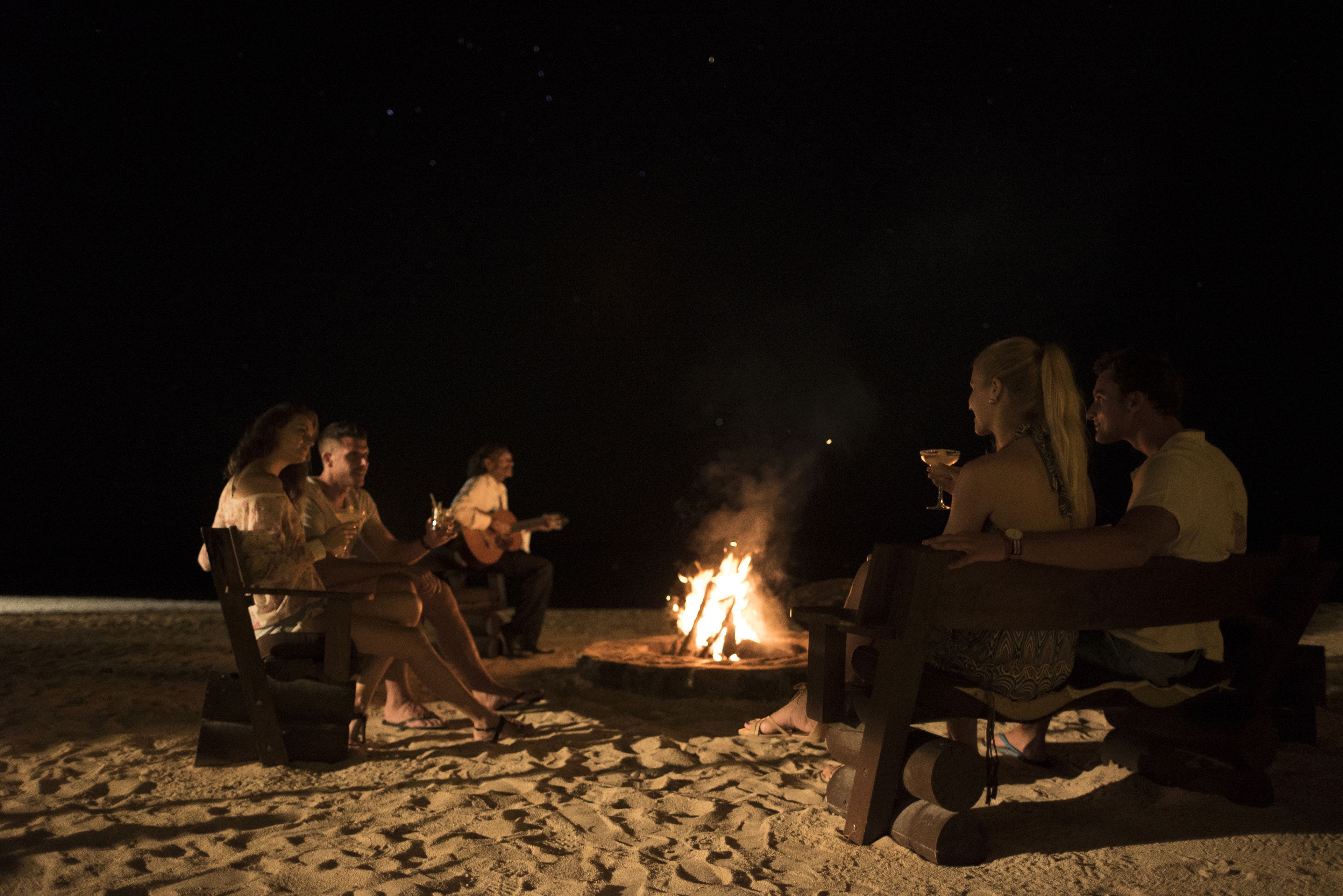 Un grupo de personas sentadas alrededor de una fogata en una playa arenosa de noche.