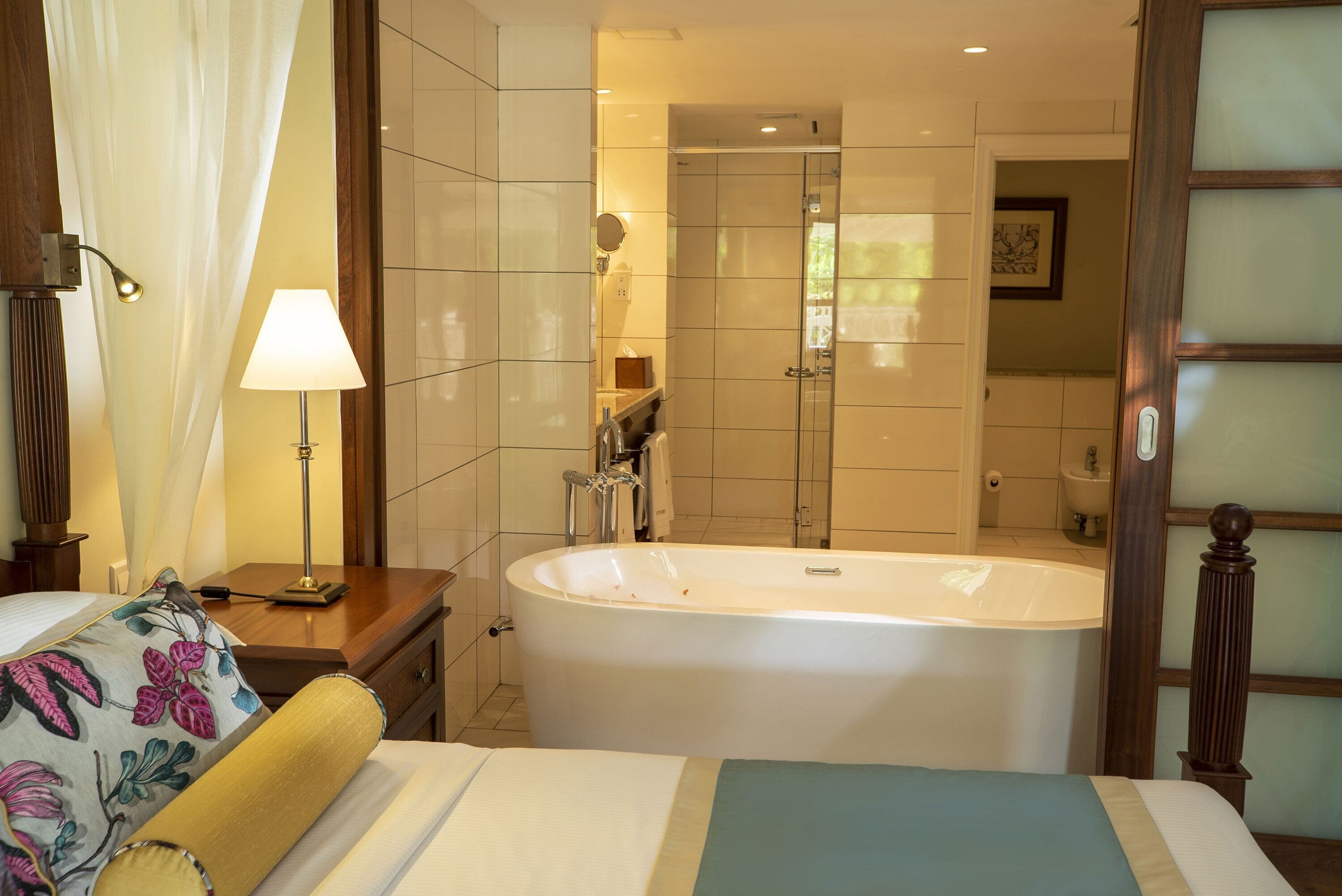 Habitación de hotel con una bañera grande, cama con ropa de cama floral, mesita de noche con lámpara y puerta de madera con paneles de vidrio texturizado.