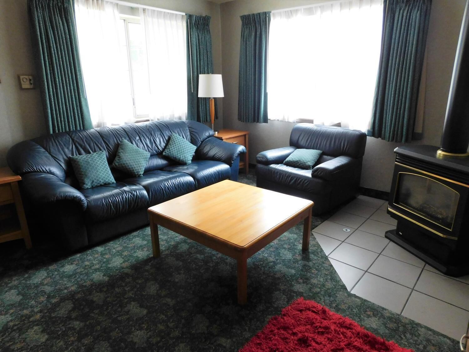 A living room with a dark leather sofa, a wooden coffee table, and a red rug. There is a black fireplace and a lamp on a side table.