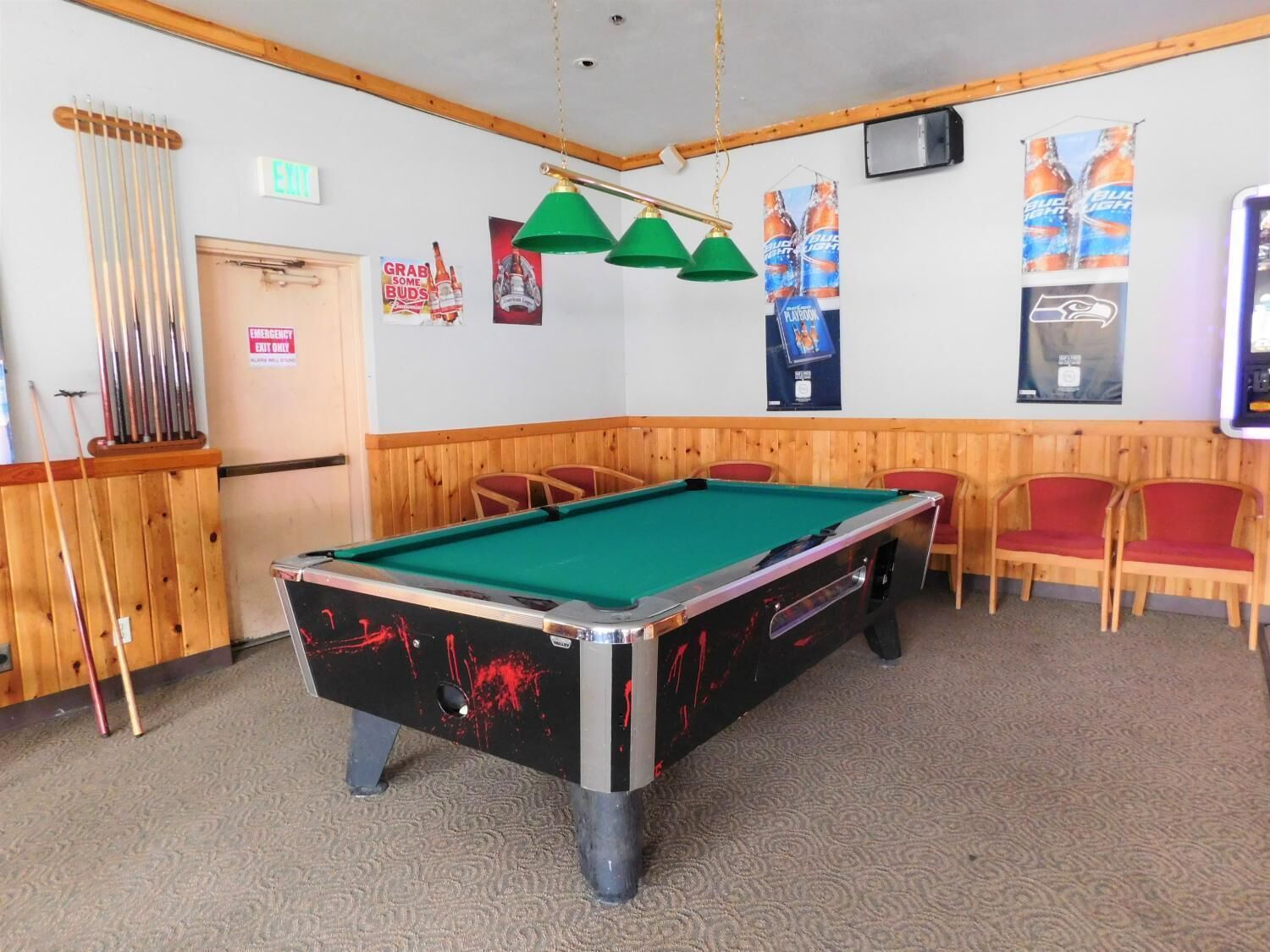 A pool table with green felt, surrounded by wooden chairs and a pool cue rack.