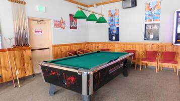 A pool table with green felt, surrounded by wooden chairs and a pool cue rack.
