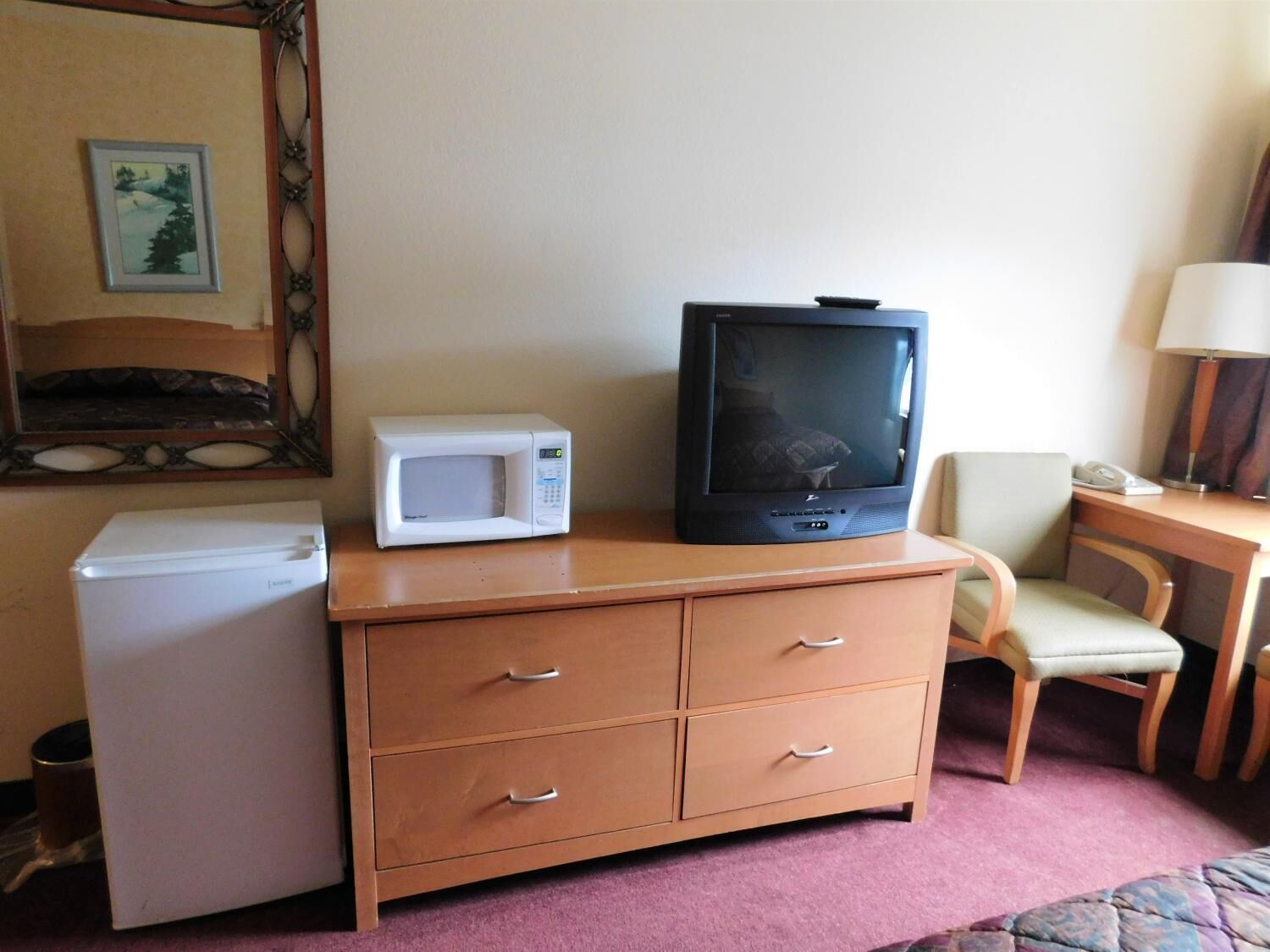 Quarto de hotel compacto com penteadeira de madeira, micro-ondas, televisão, pequena geladeira, uma cadeira e escrivaninha com luminária.
