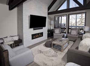 Una sala de estar moderna con televisión de pantalla plana, chimenea, asientos cómodos y ventanas grandes que ofrecen vistas al exterior.
